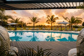 Ein Swimmingpool mit Liegestühlen und Sonnenschirmen, umgeben von Palmen, unter einem Sonnenuntergangshimmel.