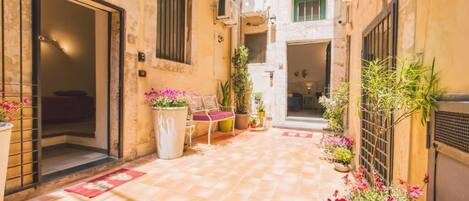 Une cour étroite au sol carrelé, un espace aménagé avec des chaises, des plantes en pot et une porte donnant sur un espace intérieur.
