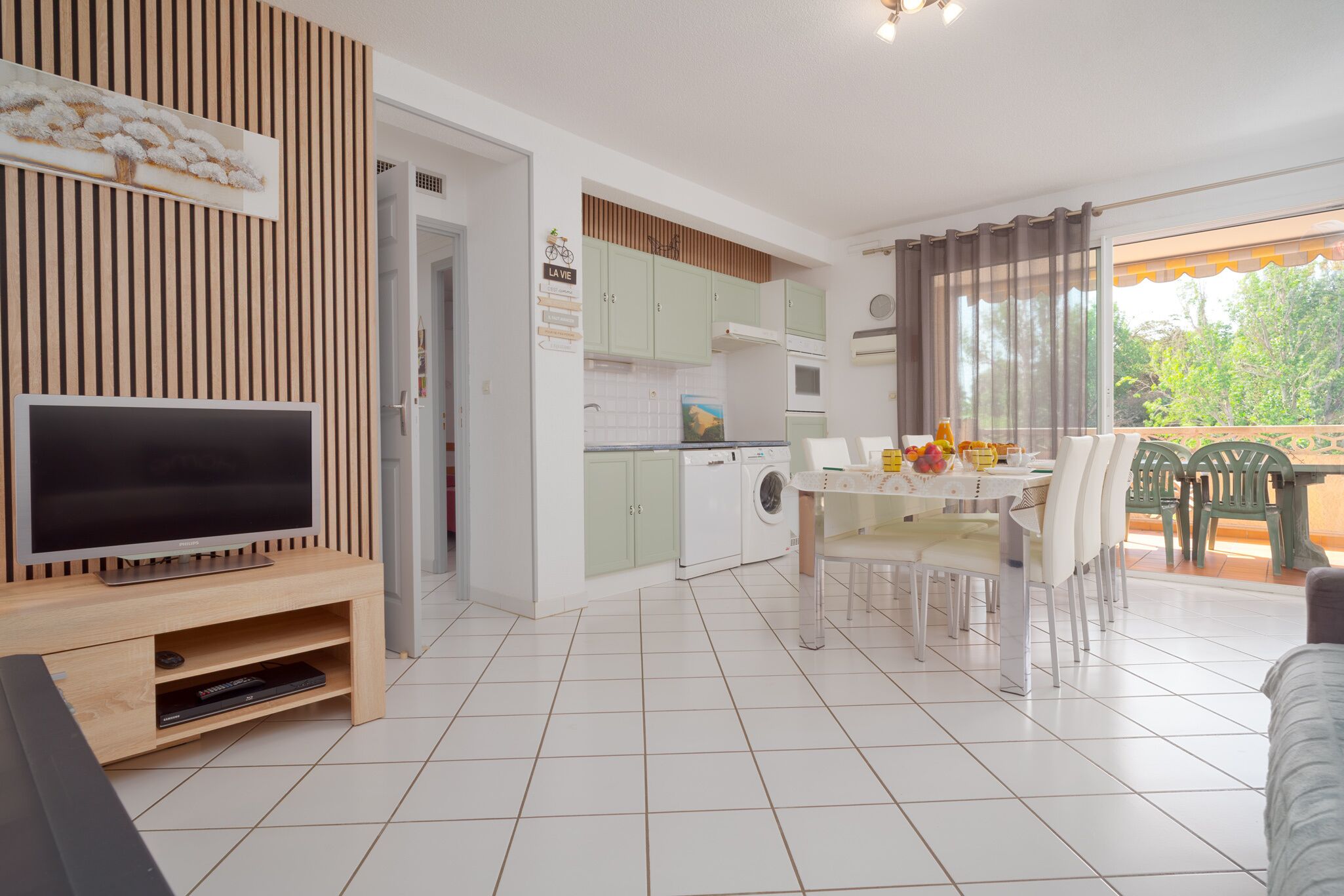 Ein modernes Wohnzimmer mit Flachbildfernseher, einer Essecke mit Tisch für zwei Personen und einer Küche mit Waschmaschine.