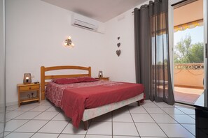 A bedroom with a bed, bedside tables, an air conditioning unit, and a view through a window with curtains. - T2 climatisé avec grande terrasse – Plage et Port à pied (Saint-Cyprien)