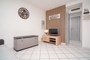A modern living room with a flat-screen TV, wooden entertainment unit, and a decorative wall clock. - T2 climatisé avec grande terrasse – Plage et Port à pied (Saint-Cyprien)