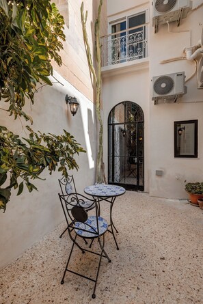 Une cour avec une table et une chaise, un pot de fleurs et une porte surmontée d’un portail en fer forgé.