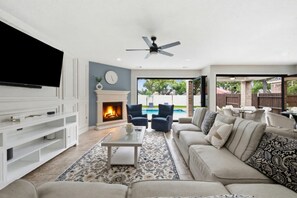 A living room with a fireplace, a television, a ceiling fan, and a view of a backyard with a pool. - The Grand Sunnyview Home: Pool & Hot Tub / Movie Room (Houston)