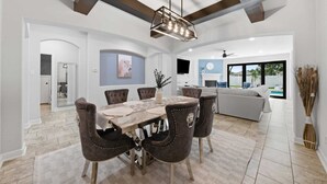 Un comedor moderno con una mesa de madera y sillas tapizadas, un techo con vigas a la vista, y vistas a una sala de estar con un sofá y una piscina.