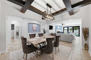 Ein stilvoller Essbereich mit Holztisch und gepolsterten Stühlen, einer Decke mit freiliegenden Balken und Blick in ein Wohnzimmer mit Sofa und Pool.
