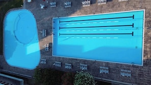 Luchtbeeld van een zwembad met meerdere banen, een apart kleiner zwembad en een terras van baksteen met buitenmeubels.
