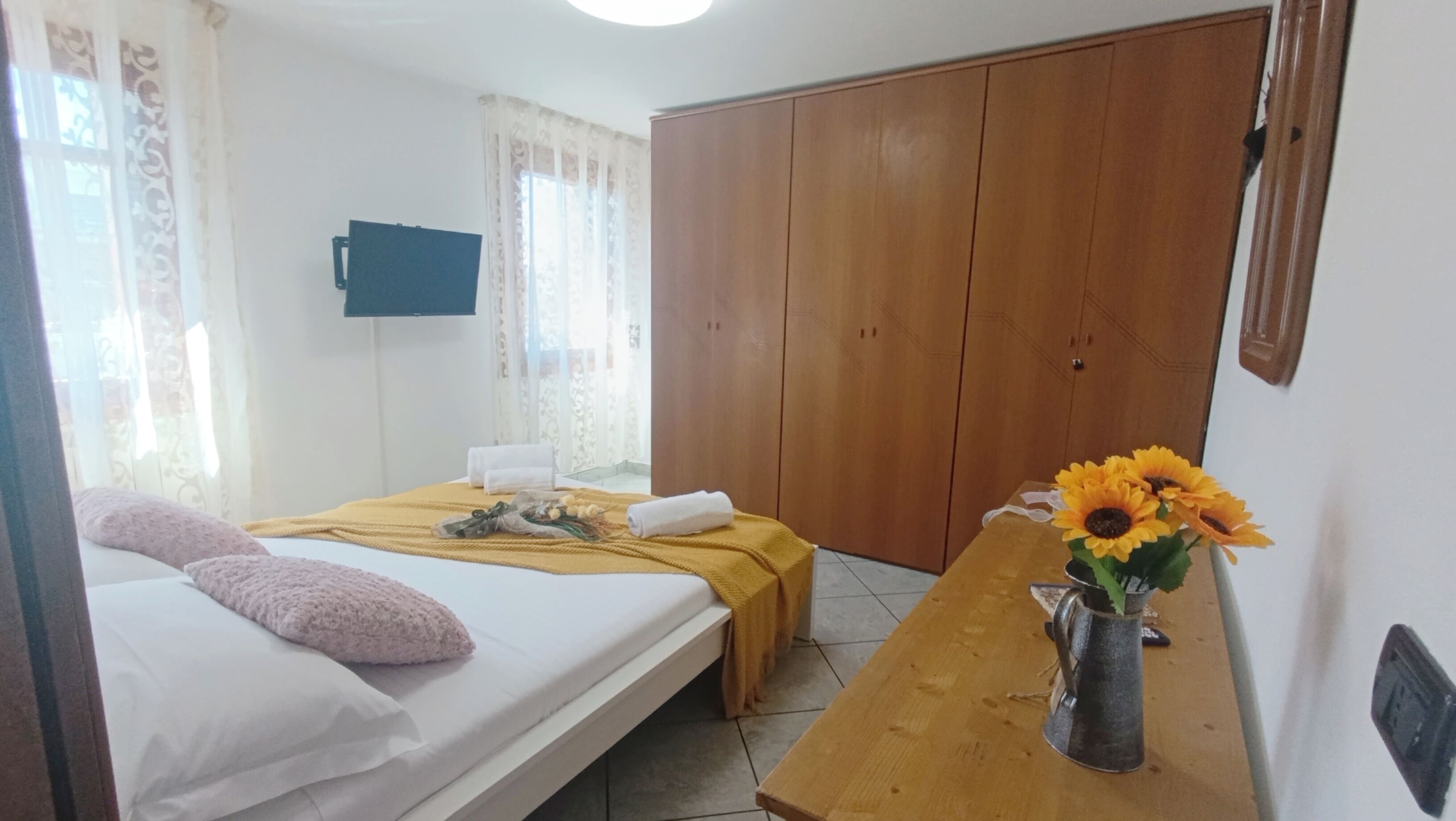 A hotel room with a bed, a wooden wardrobe, a table with a vase of sunflowers, and a television on the wall.