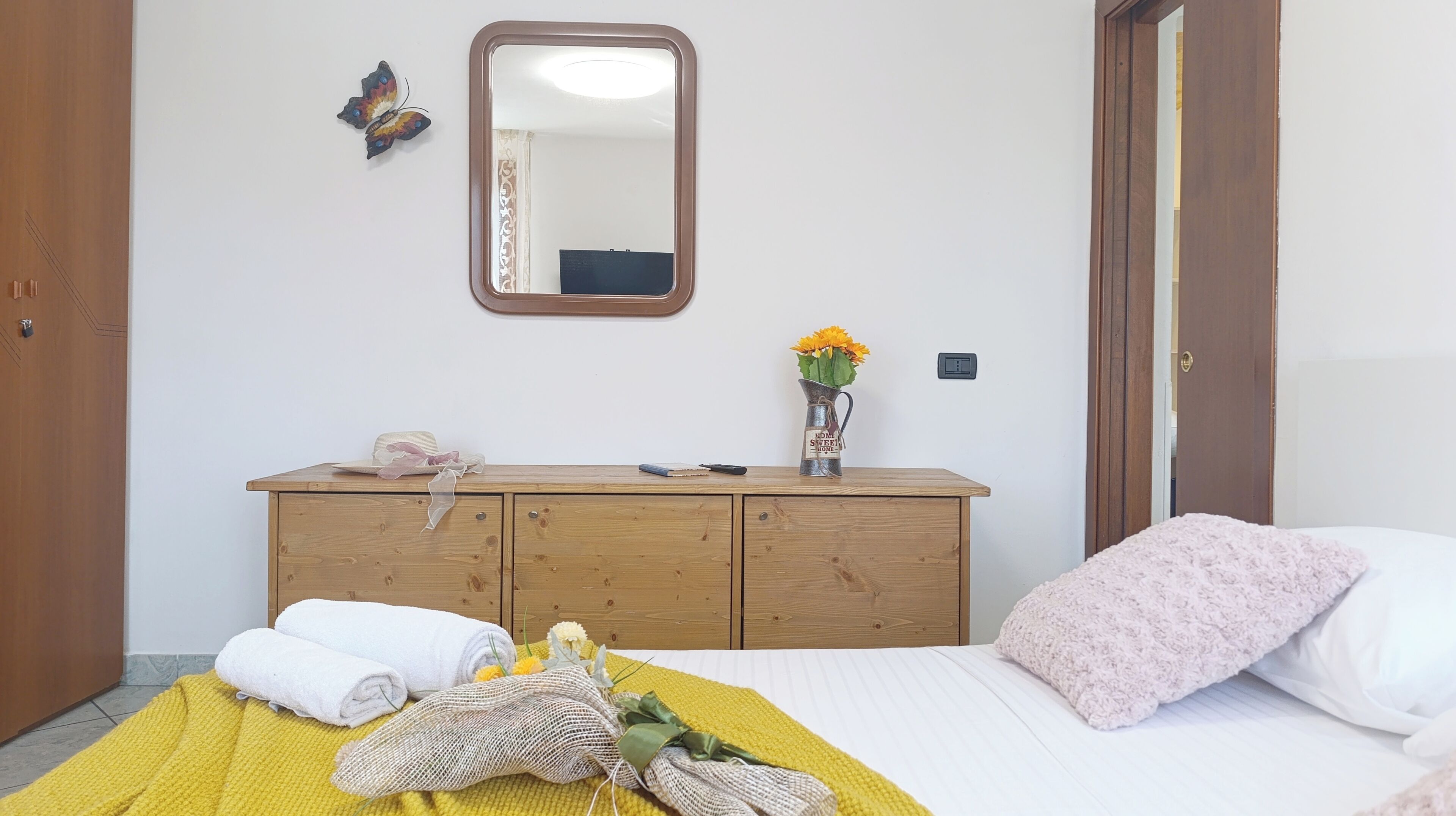 Un dormitorio con cama, cómoda de madera, espejo, un jarrón con flores y una puerta.
