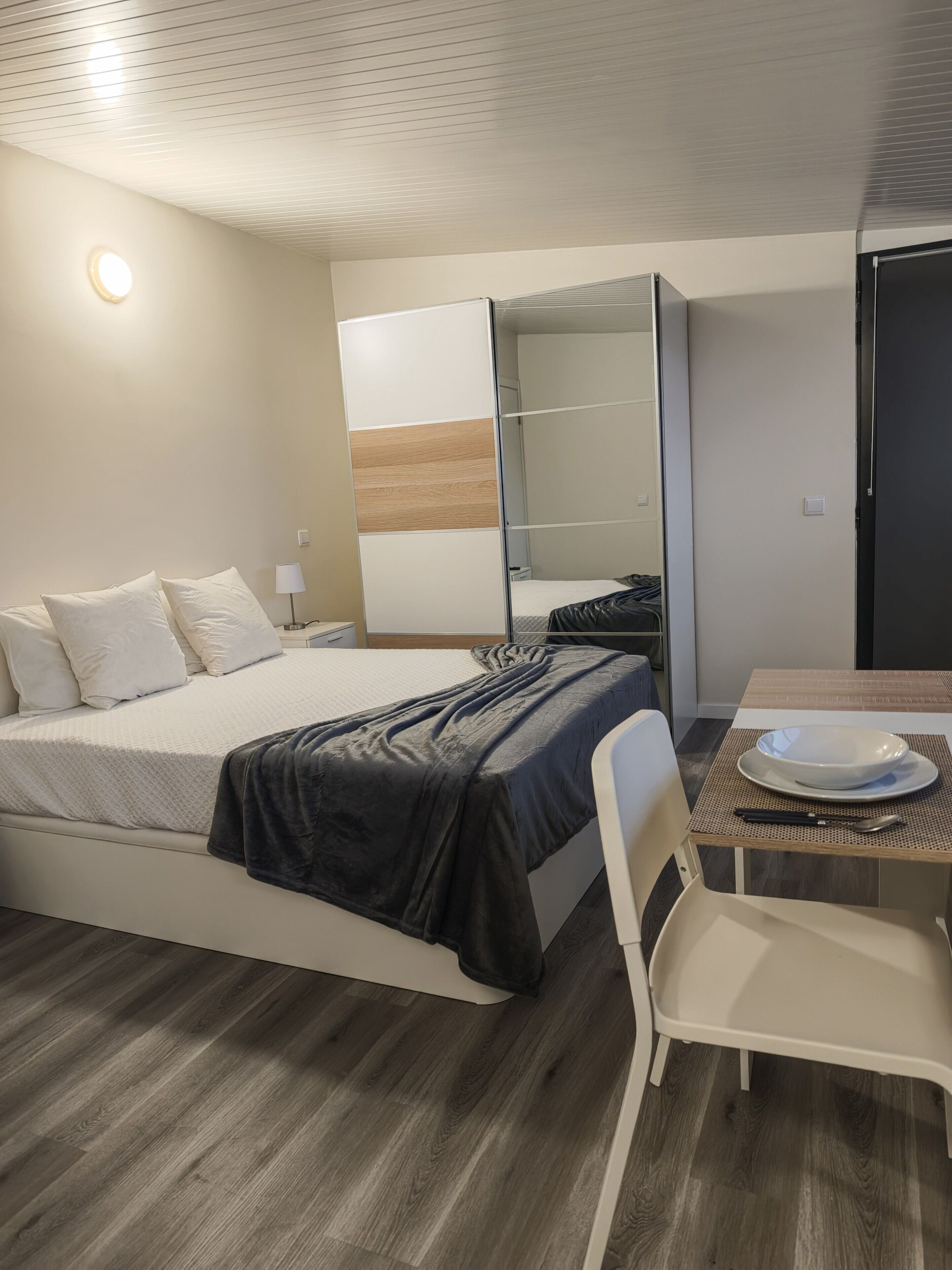 A hotel room with a bed, a desk with a chair, a mirror, and a wardrobe.