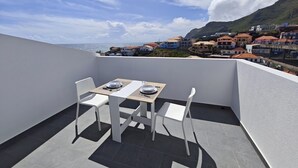 A balcony with a table and chairs, overlooking a coastal town with buildings and a mountain in the background.