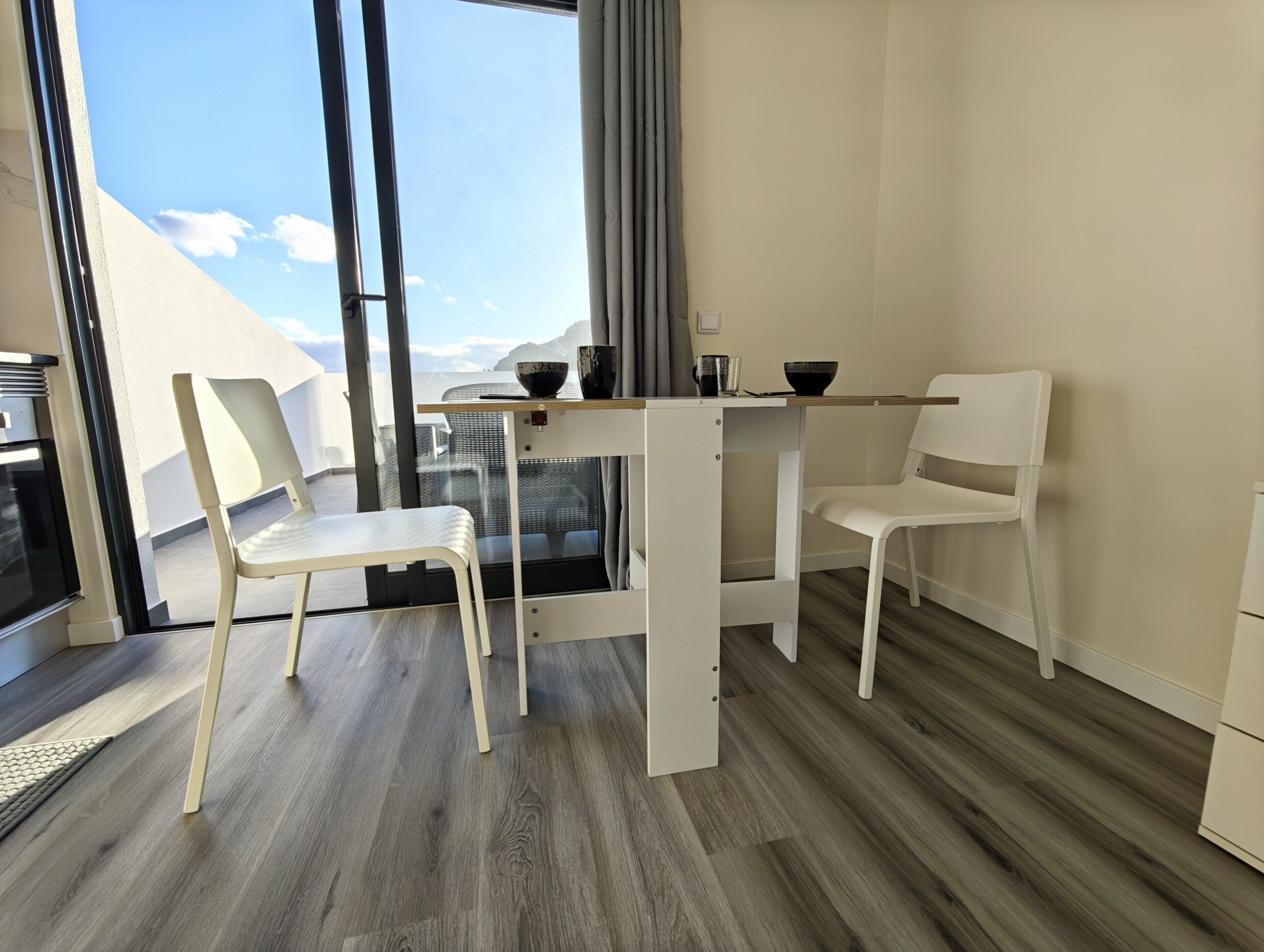 Quarto moderno com escrivaninha branca, duas cadeiras brancas, janela com cortinas e vista do céu.