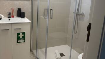 A modern bathroom with a glass-enclosed shower, a sink with a mirror above, and a sign indicating first aid availability.