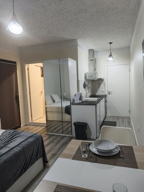 A compact hotel room with a bed, a small kitchenette, a mirror, and a dining area with a table set for two.