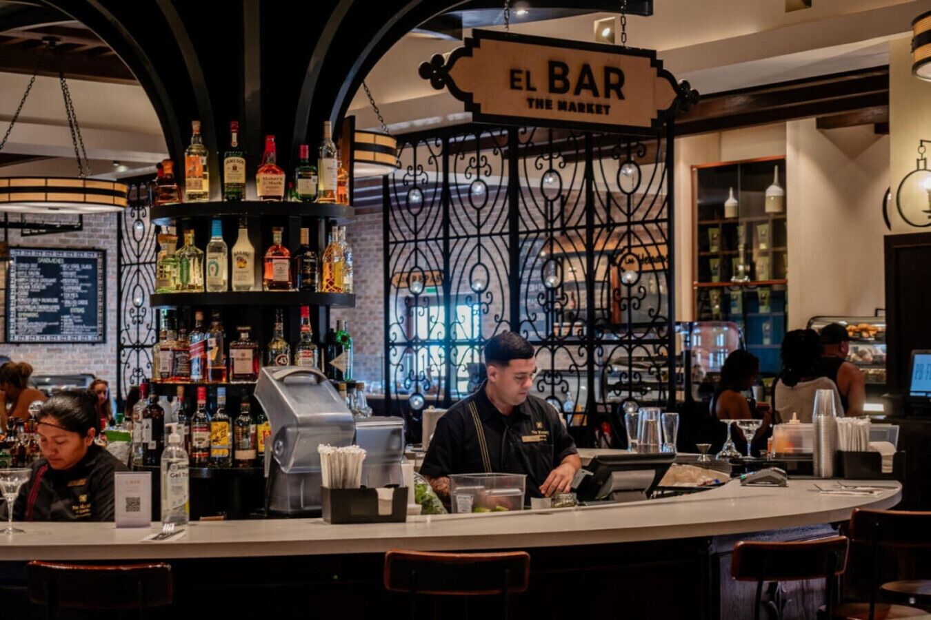 A bar area with a sign reading 'EL BAR THE MARKET,' shelves of liquor bottles, a cash register, and bar stools.