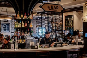Un espace bar avec une enseigne indiquant « EL BAR THE MARKET », des étagères remplies de bouteilles d’alcool, une caisse enregistreuse et des tabourets de bar.