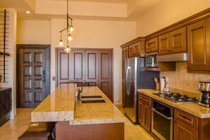 A kitchen with a central island, stainless steel appliances, wooden cabinets, and a pendant light fixture. - Quivira Luxury Penthouse | Ocean Views & Resort Amenities (Cabo San Lucas)