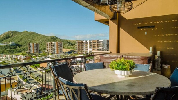 A balcony with a round table and chairs, a potted plant, and a view of buildings and hills. - Quivira Luxury Penthouse | Ocean Views & Resort Amenities (Cabo San Lucas)