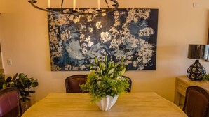 A dining area with a wooden table, chairs, a floral centerpiece, and a large abstract painting on the wall. - Quivira Luxury Penthouse | Ocean Views & Resort Amenities (Cabo San Lucas)