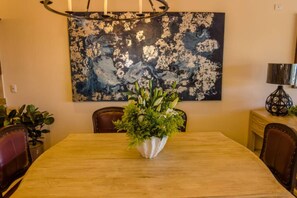 A dining area with a wooden table, chairs, a floral centerpiece, and a large abstract painting on the wall. - Quivira Luxury Penthouse | Ocean Views & Resort Amenities (Cabo San Lucas)