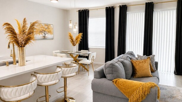 Ein modernes Wohnzimmer mit grauem Sofa, einem Couchtisch und einem Essbereich mit weißem Tisch und goldenen Stühlen.