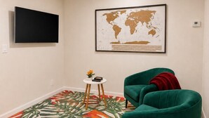 Ein Zimmer mit einem an der Wand befestigten Flachbildfernseher, einer Weltkarte als Wandbild über einem grünen Sessel und einem kleinen runden Tisch mit Pflanze und Buch.