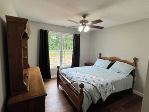Ein Schlafzimmer mit einem hölzernen Bett, einem Nachttisch, einer Kommode, einem Deckenventilator und einem Fenster mit Vorhängen.