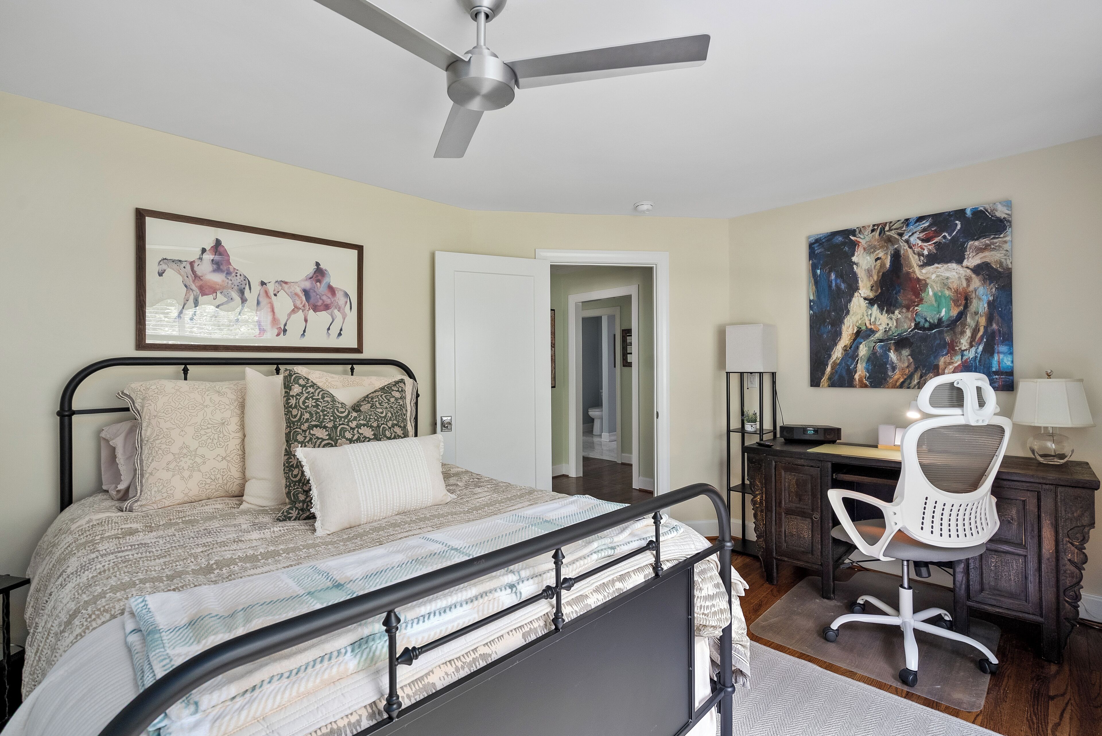 Un dormitorio con cama, escritorio, silla, ventilador de techo y obras de arte en las paredes.