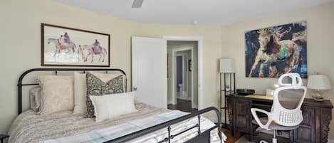 Un dormitorio con cama, escritorio, silla, ventilador de techo y obras de arte en las paredes.