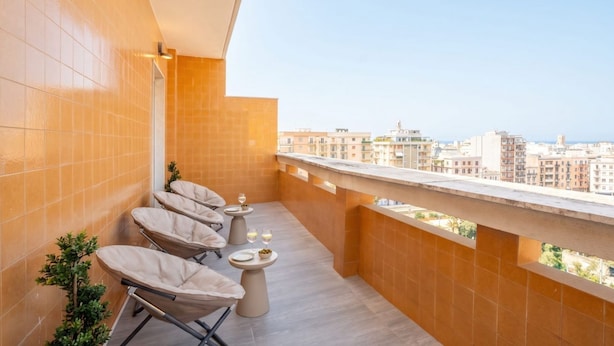 Ein Balkon mit vier gepolsterten Stühlen, zwei kleinen runden Tischen mit Getränken und Blick auf eine Stadtlandschaft.