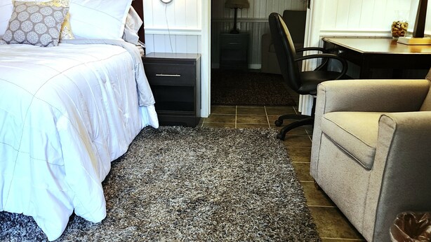 Quarto de hotel com cama, mesa de cabeceira, luminária, cadeira, escrivaninha e tapete.