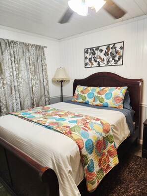 A bedroom with a wooden bed, a patterned bedspread, a wall-mounted picture, and a floor lamp.
