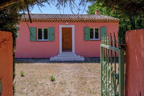 Un bâtiment rose avec une porte en bois et des volets verts, un petit escalier menant  à l’entrée et un portail en métal au premier plan.