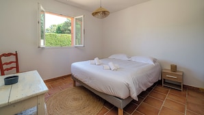 Ein Schlafzimmer mit Bett, Sessel, Tisch, Nachttisch und Fenster mit Blick auf Grünflächen.