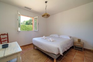 A bedroom with a bed, a chair, a table, a bedside table, and a window with a view of greenery. - Maison Vigneron | Domaine Tour Campanets (Le Puy-Sainte-Réparade)