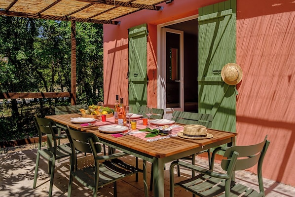 An outdoor dining area with a wooden table, green chairs, and a variety of fruits and drinks. - Maison Vigneron | Domaine Tour Campanets (Le Puy-Sainte-Réparade)