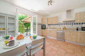 A kitchen with a dining area, open cabinets, a dishwasher, and a view of the outdoors. - Maison Vigneron | Domaine Tour Campanets (Le Puy-Sainte-Réparade)