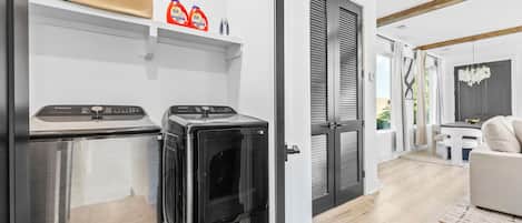 A modern laundry room with stainless steel appliances, a white wall, and a wooden floor.