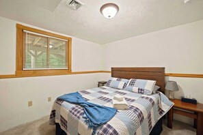 A bedroom with a bed, a nightstand, a lamp, and a window with blinds. - Family Cabin Near Story Land & Attitash (Bartlett)