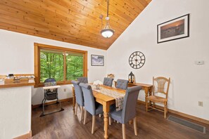 A dining area with a wooden table, grey upholstered chairs, a wooden ceiling, a large window with a view of trees, and a wall clock. - Family Cabin Near Story Land & Attitash (Bartlett)