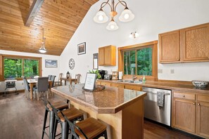 A modern kitchen with a central island, wooden cabinets, and a view of the outdoors through large windows. - Family Cabin Near Story Land & Attitash (Bartlett)