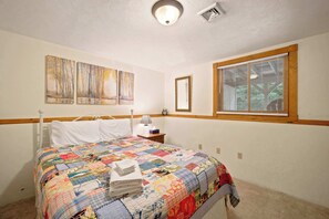 A bedroom with a bed, a nightstand, a lamp, a window with blinds, and wall art. - Family Cabin Near Story Land & Attitash (Bartlett)