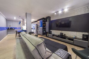 Een moderne woonkamer met een bank, een salontafel en een tv aan de muur. Achtergrond is een keuken met kasten en een magnetron.