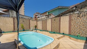 Una piscina con borde de baldosas, rodeada por un muro de piedra con alambre de púas, y un pequeño jardín con plantas en macetas.