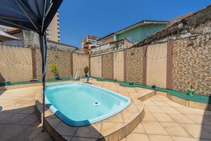 Ein Schwimmbecken mit gefliestem Sonnendeck, umgeben von einer Steinmauer mit Stacheldraht, und ein kleiner Gartenbereich mit Topfpflanzen.