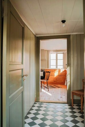 A hallway with a checkered floor leading to a kitchen area with wooden cabinets and a window. - Authentic 1800s cottage in Småland (Svenstorp)
