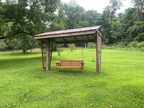A wooden gazebo with a swing, surrounded by a grassy area and trees.