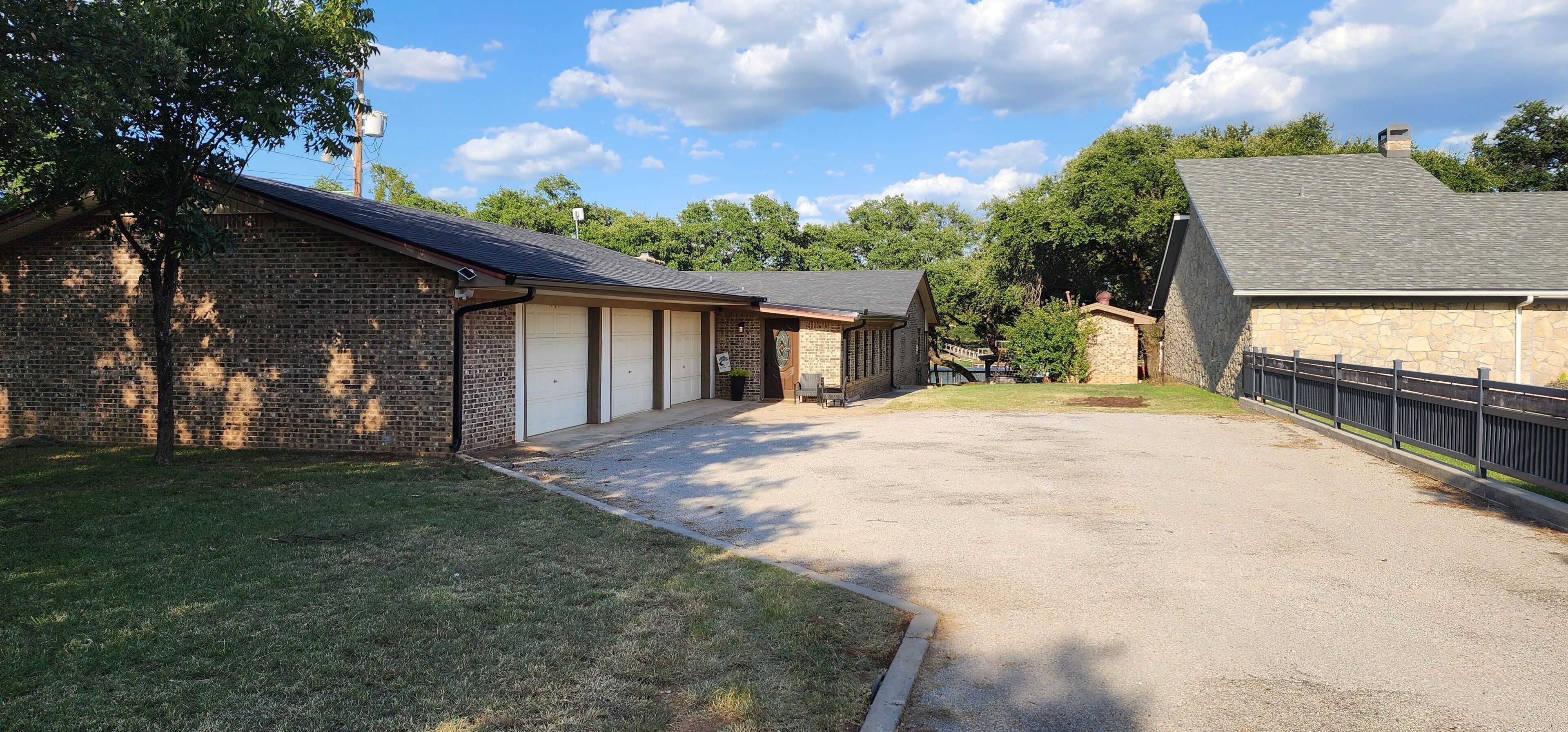 A property with a gravel driveway, a brick garage, and a stone building with a metal fence.