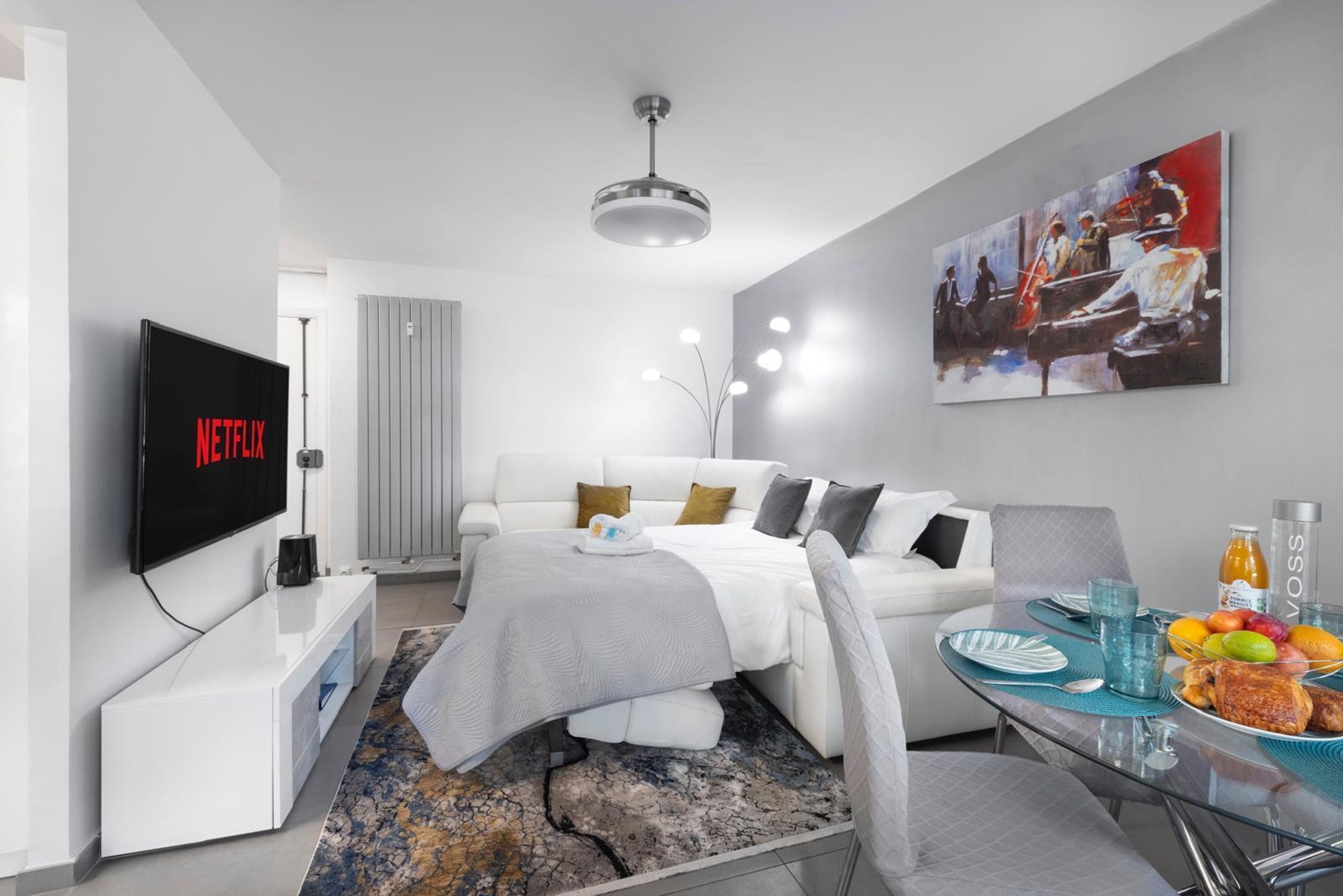 A modern hotel room with a bed, a TV displaying the Netflix logo, a dining area with a table set for two, and a painting on the wall.