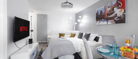 A modern hotel room with a bed, a TV displaying the Netflix logo, a dining area with a table set for two, and a painting on the wall.