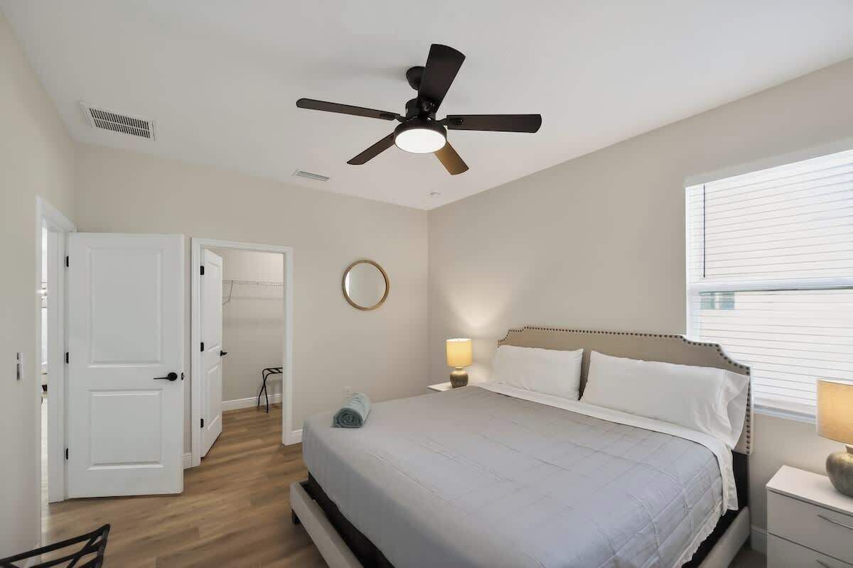 A bedroom with a bed, a ceiling fan, a bedside table with a lamp, a window with blinds, and a door leading to another room.
